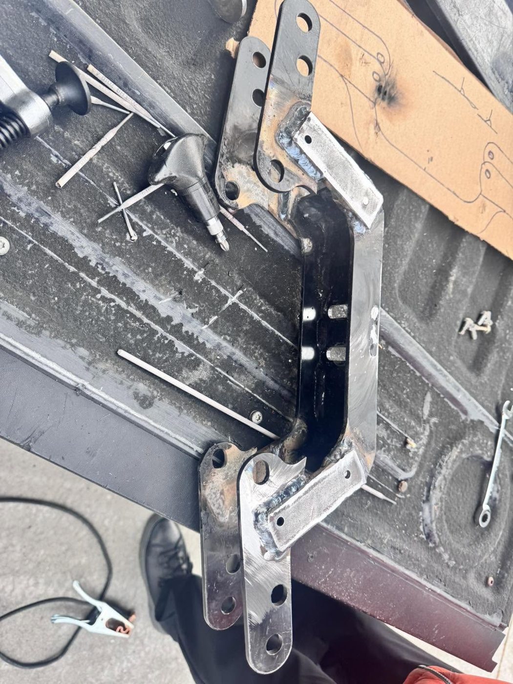 Two steel brackets clamped and tack-welded together on a steel work surface, with tools nearby