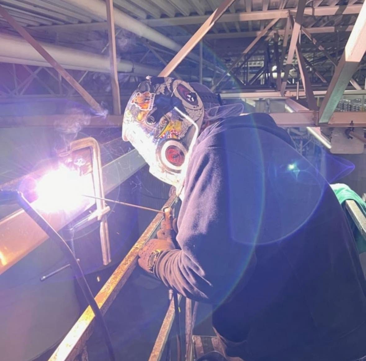 Welder in a hood welding overhead on structural steel inside a commercial building, with a bright arc