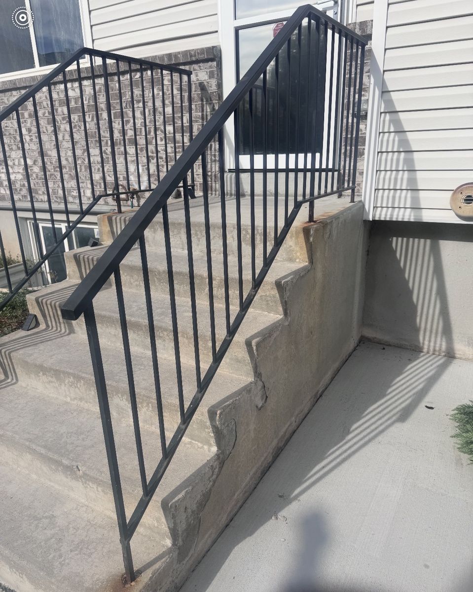 Old black steel railing on concrete entry steps, with rust at the post bases