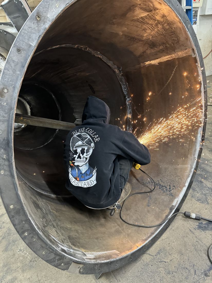 A welder in a black hoodie crouched inside a large steel cone, grinding a seam with sparks flying